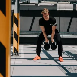 Młody mężczyzna w czarnym stroju sportowym i pomarańczowych butach ćwiczy z dwoma odważnikami kettlebell na parkingu, oświetlony promieniami słońca padającymi przez metalową konstrukcję.