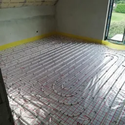 Białe rury ogrzewania podłogowego rozłożone na folii z czerwoną kratką w pokoju na poddaszu z oknem i izolacją na skosach.