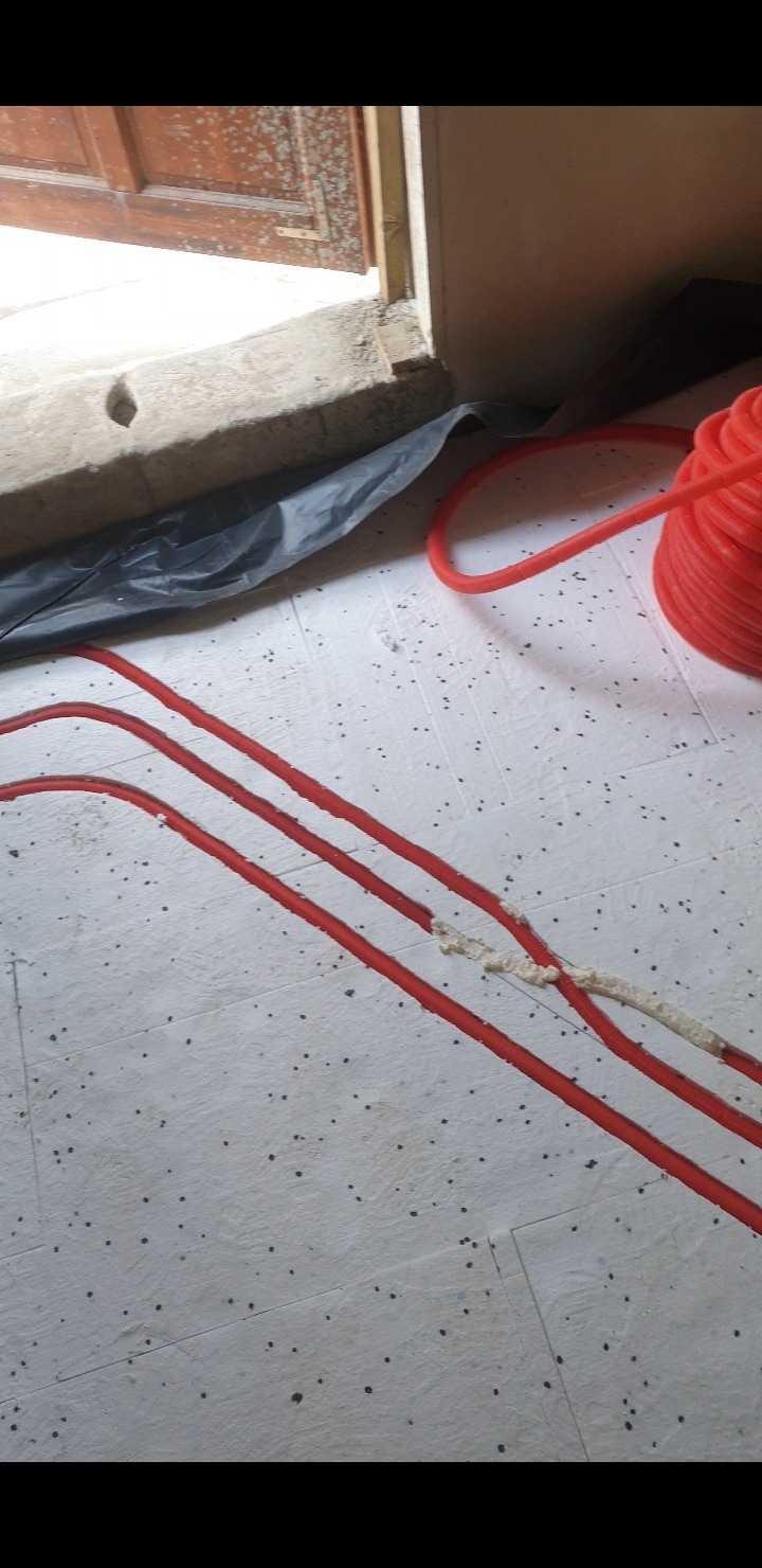 Czerwone rurki ogrzewania podłogowego przytwierdzone pianką montażową do białej płyty izolacyjnej z czarnymi kropkami, widoczny fragment drzwi i folii ochronnej.