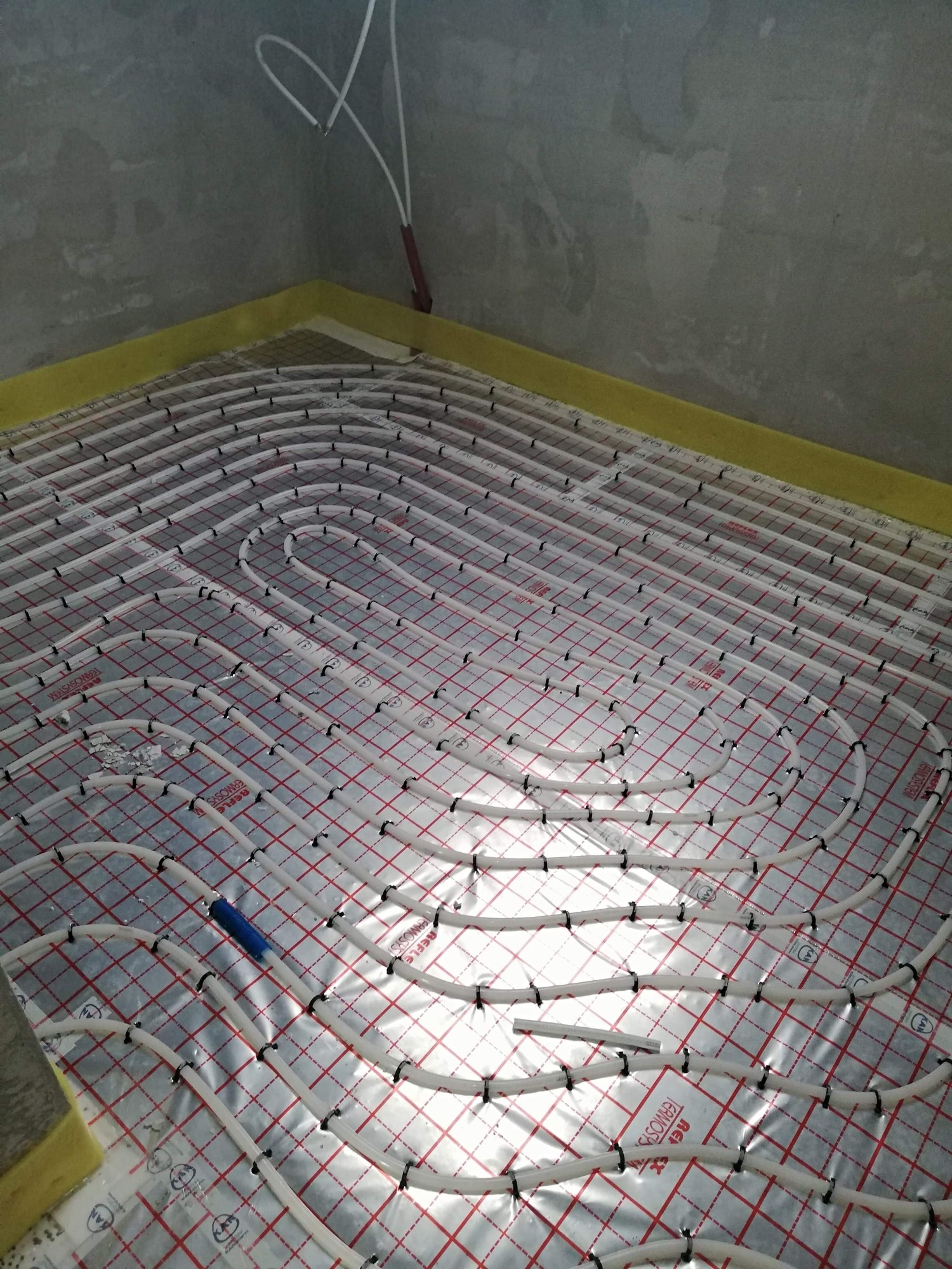 Białe rury ogrzewania podłogowego rozłożone na srebrnej folii z czerwoną kratką, zamocowane czarnymi klipsami, widoczne fragmenty żółtej pianki przy ścianach.