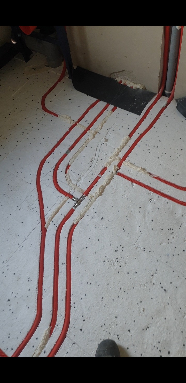 Czerwone rury ogrzewania podłogowego ułożone w bruzdach w podłodze, zabezpieczone pianką montażową, widoczny fragment buta.
