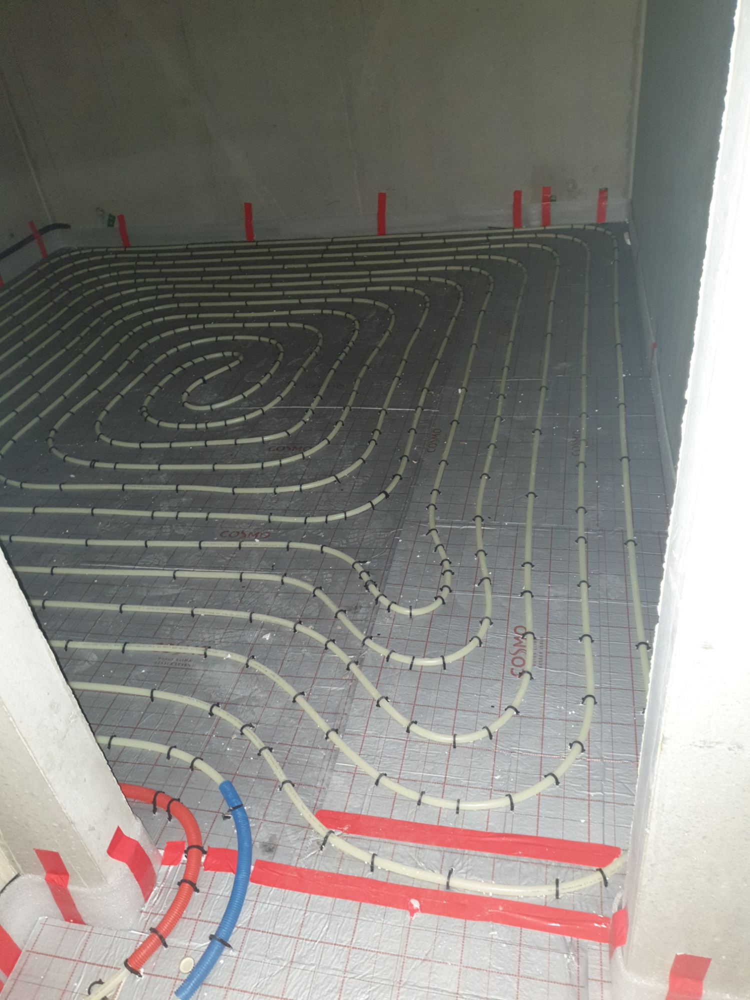 Rozłożone rury ogrzewania podłogowego na izolacji z siatką, mocowane plastikowymi uchwytami, czerwona taśma na ścianach, niebieska i pomarańczowa rura w rogu.