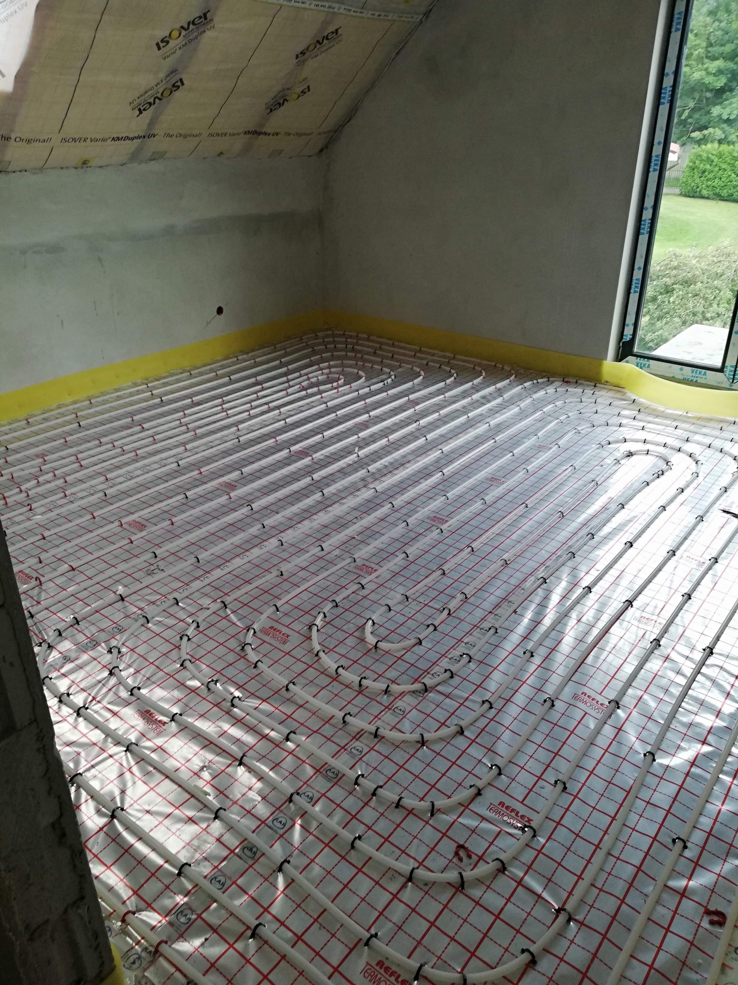 Białe rury ogrzewania podłogowego rozłożone na folii z czerwoną kratką w pokoju na poddaszu z oknem i izolacją na skosach.