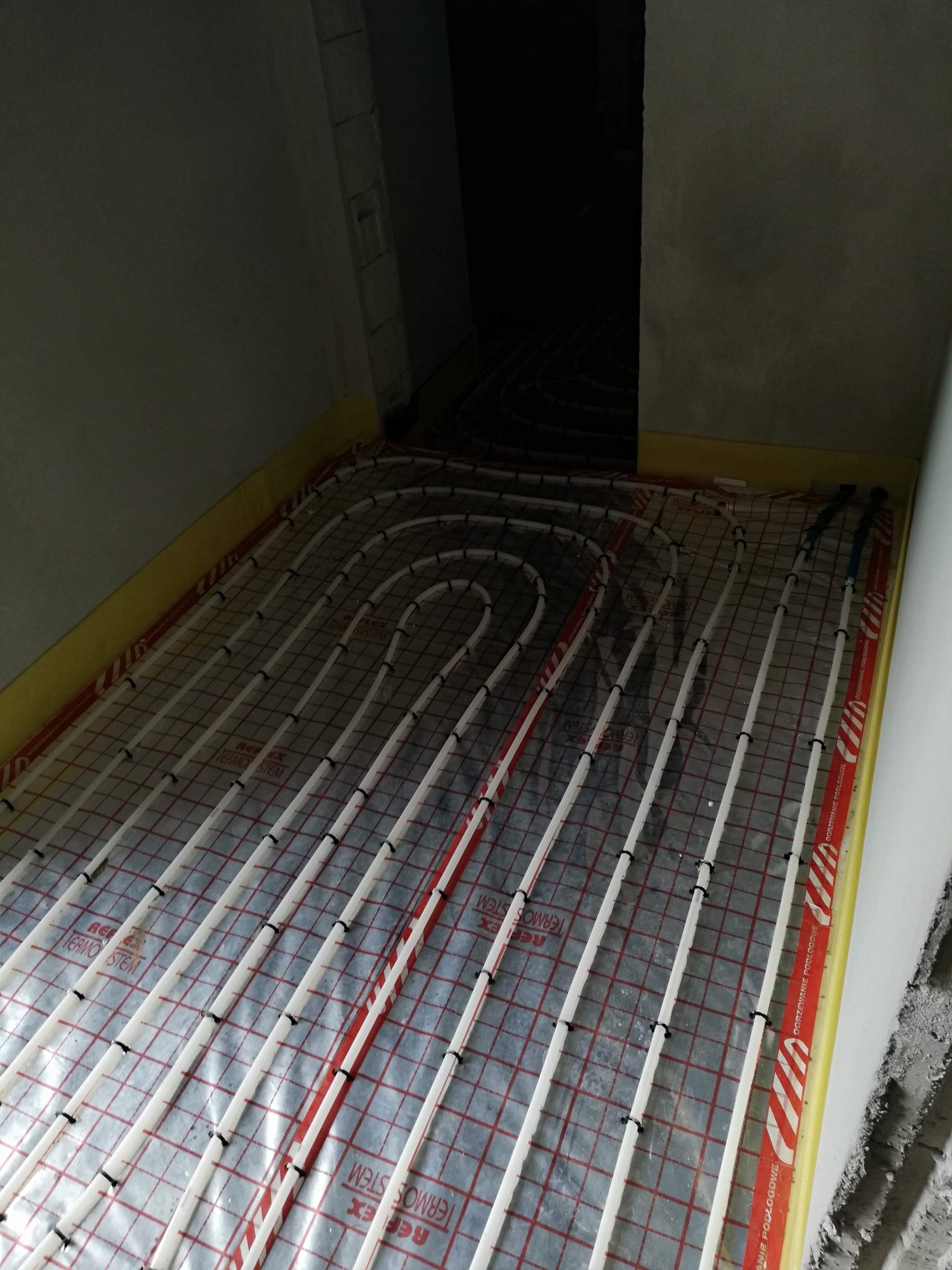 Rozłożona instalacja ogrzewania podłogowego na folii z nadrukiem, widoczne rurki mocowane czarnymi uchwytami, żółta taśma brzegowa przy ścianie w surowym stanie.