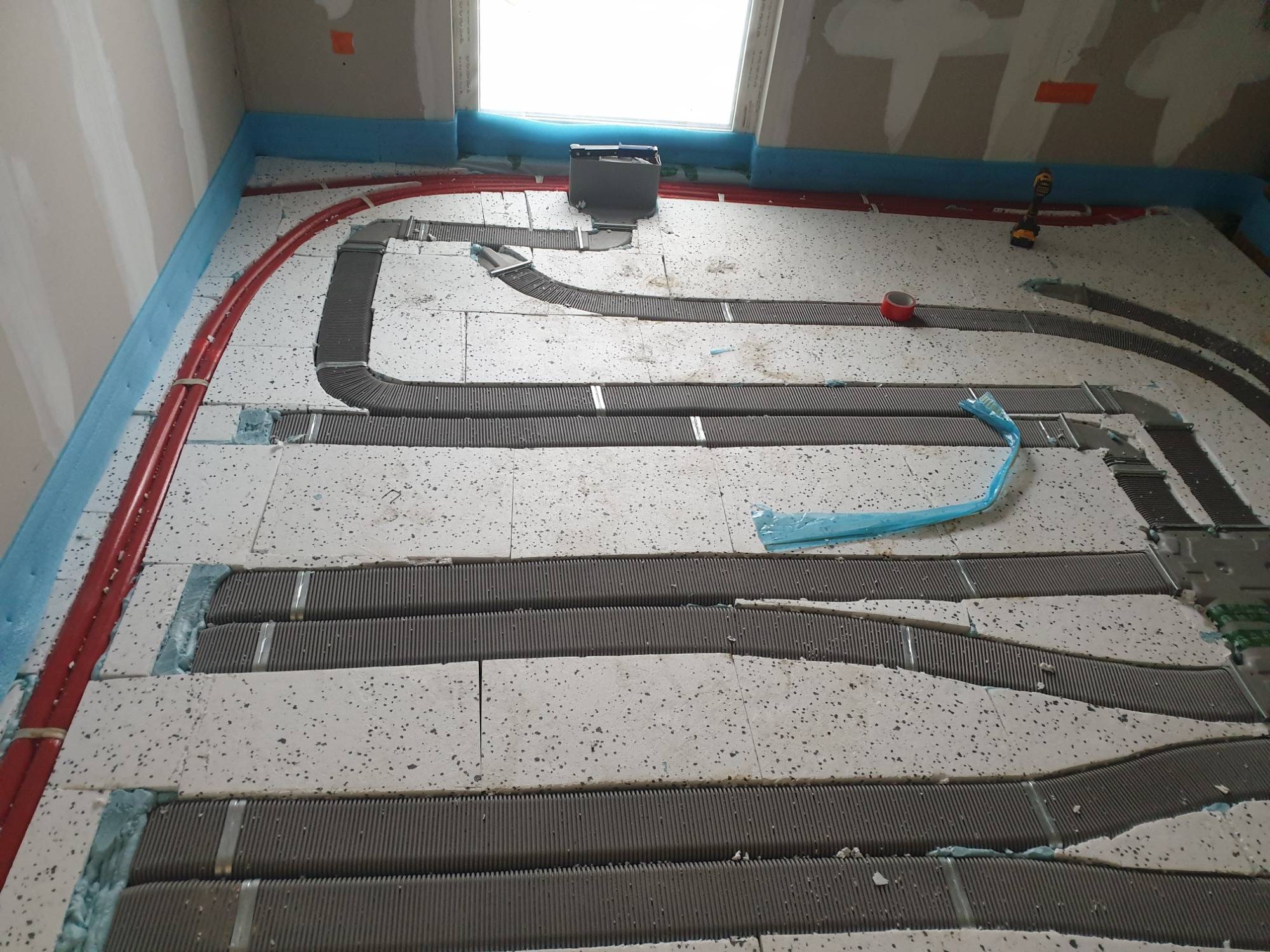 Niedokończona instalacja ogrzewania podłogowego: czerwone rury i szare panele grzewcze ułożone na białym styropianie, widoczna wiertarka akumulatorowa, okno dachowe w tle.