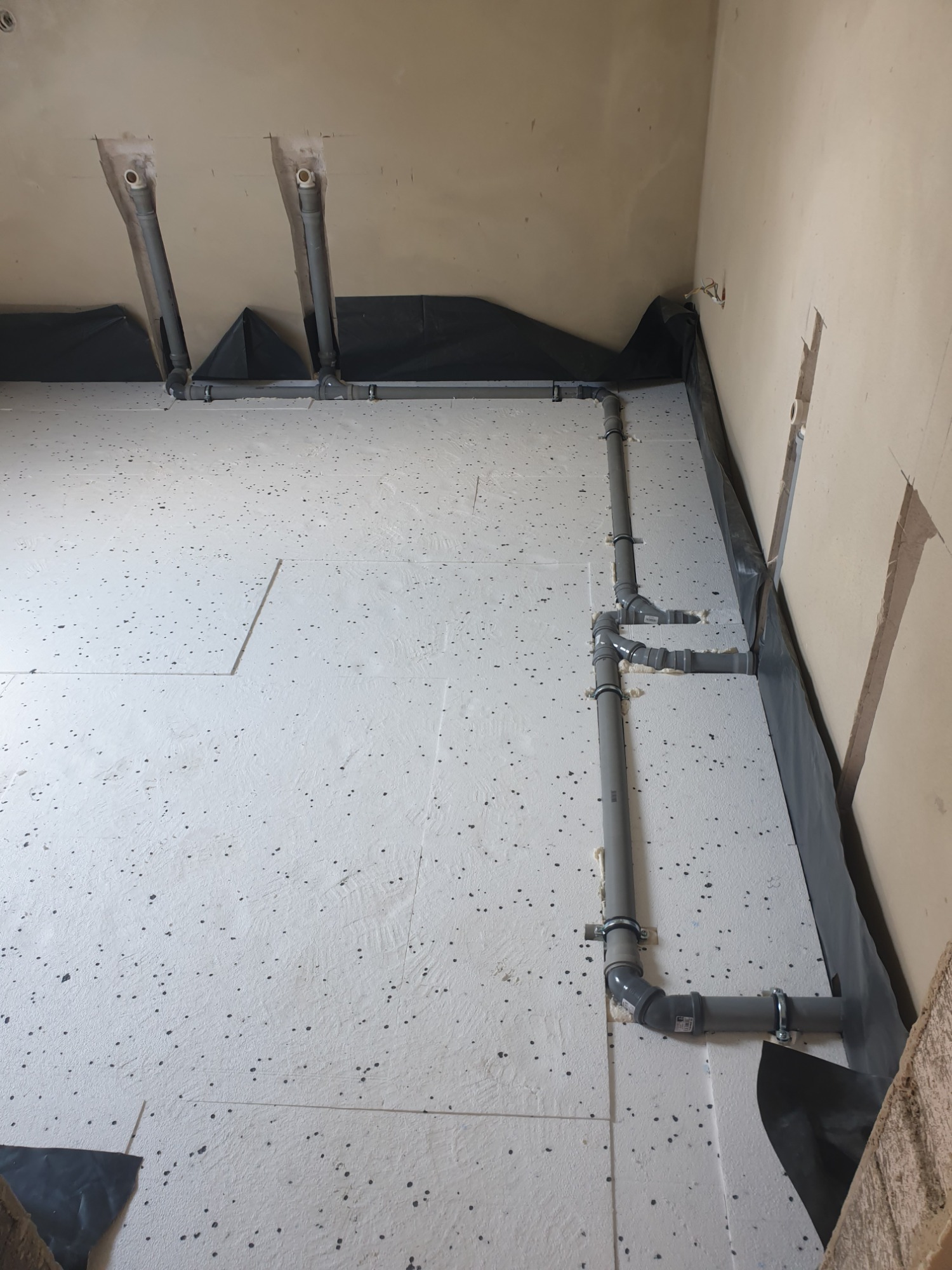 Szare rury kanalizacyjne zamontowane na białej powierzchni pokrytej czarnymi plamkami, zabezpieczone czarną folią przy ścianie. Widoczne mocowania rur do podłoża.