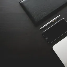 Czarny skórzany notatnik, srebrny długopis, smartfon i fragment laptopa na ciemnym drewnianym biurku z widoczną fakturą drewna.