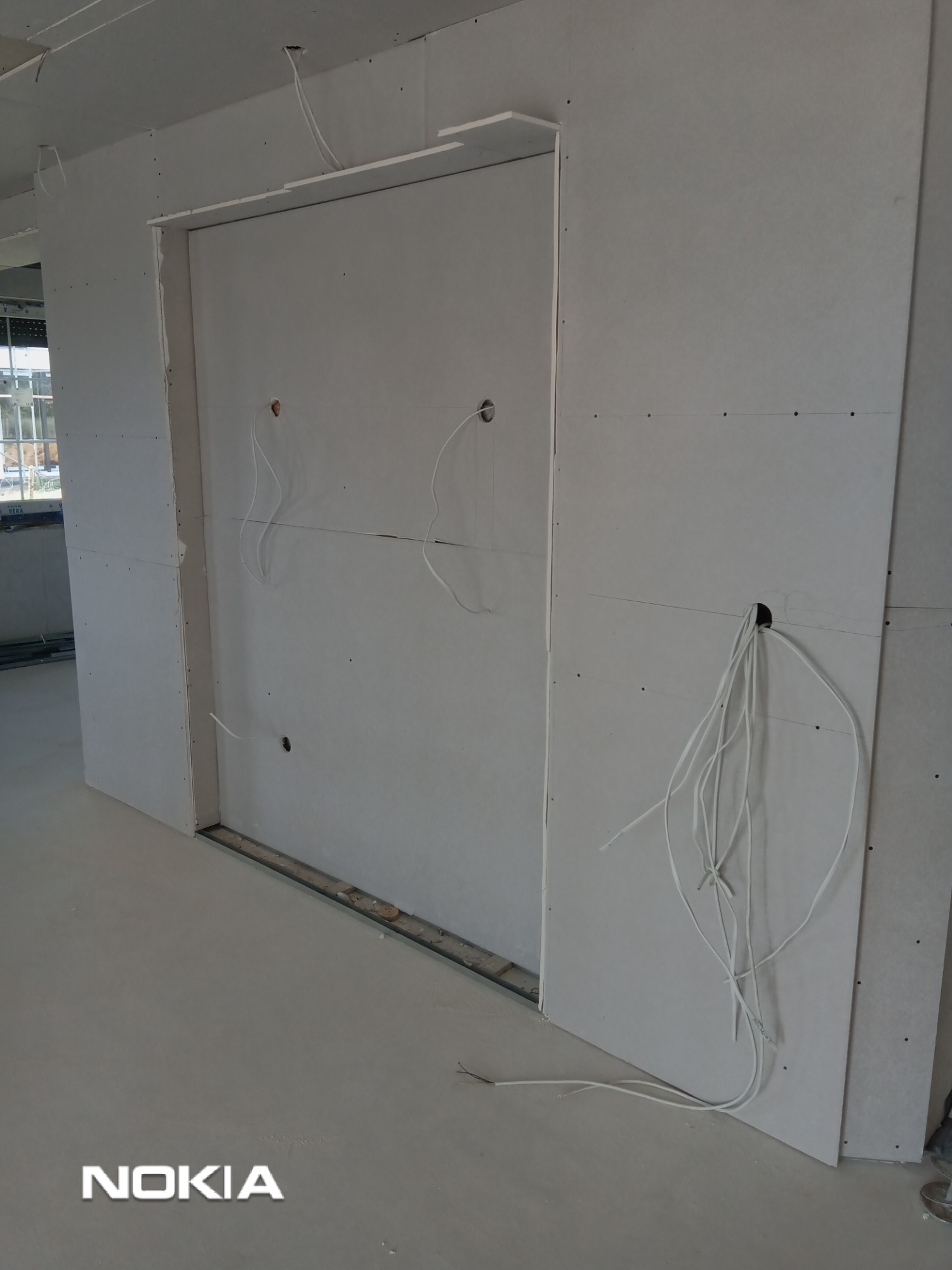 Montaż płyty karton gips w Legnicy: Widok na fragment ściany z zamontowaną płytą gipsową, widoczne przewody elektryczne wychodzące z otworów w płycie.