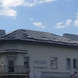 Panele fotowoltaiczne na dachu budynku z szarą dachówką, z widocznym szyldem 'Silver ul. M.Reja 23' i balkonem z kutą balustradą.
