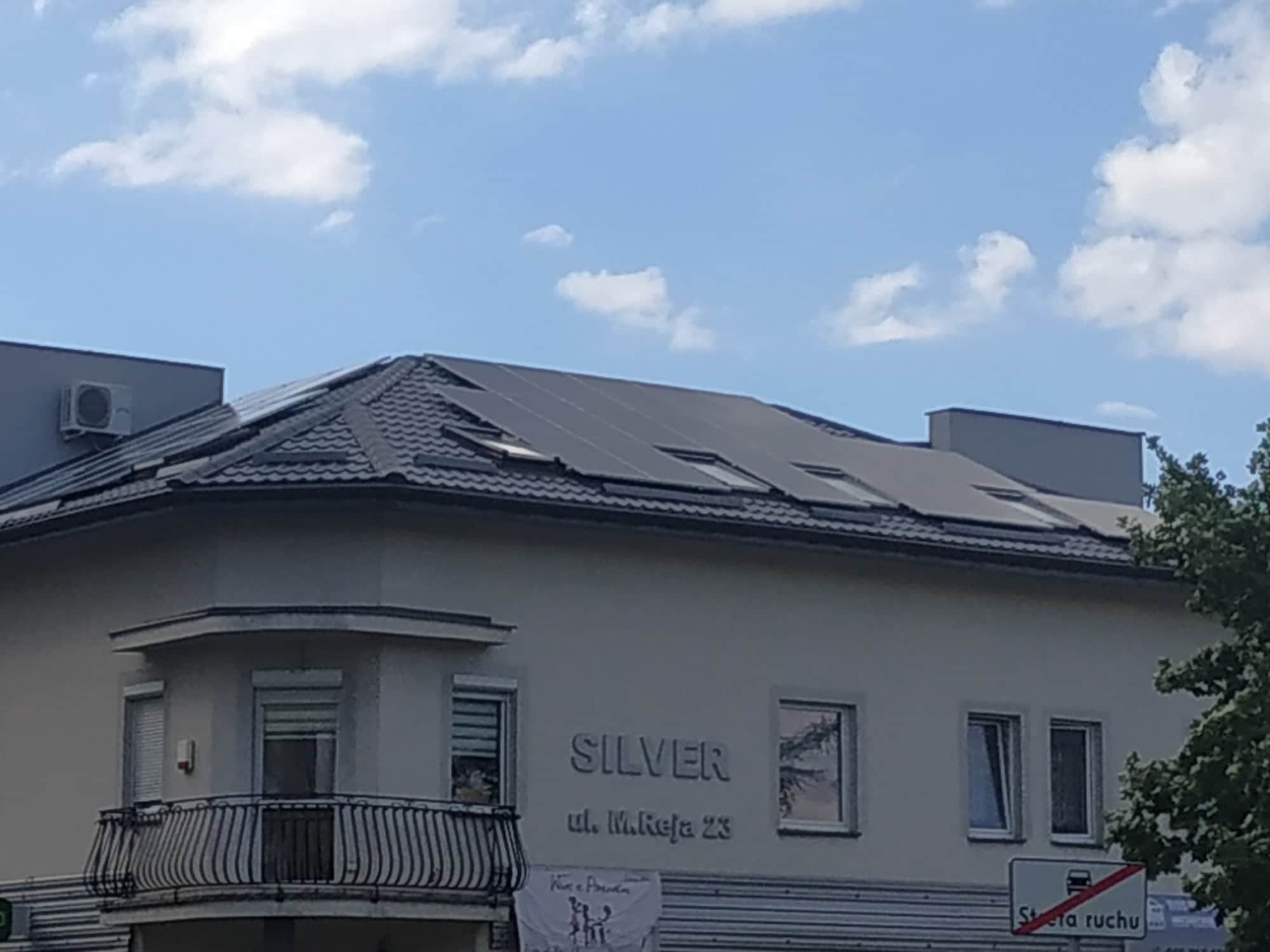 Panele fotowoltaiczne na dachu budynku z szarą dachówką, z widocznym szyldem 'Silver ul. M.Reja 23' i balkonem z kutą balustradą.
