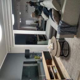 so.haus - Nowoczesny salon z szarym narożnikiem, okrągłym stolikiem kawowym z dekoracyjnymi świecami, oraz szafką RTV z rośliną w doniczce, widok z boku, z odbiciem osoby w oknie.