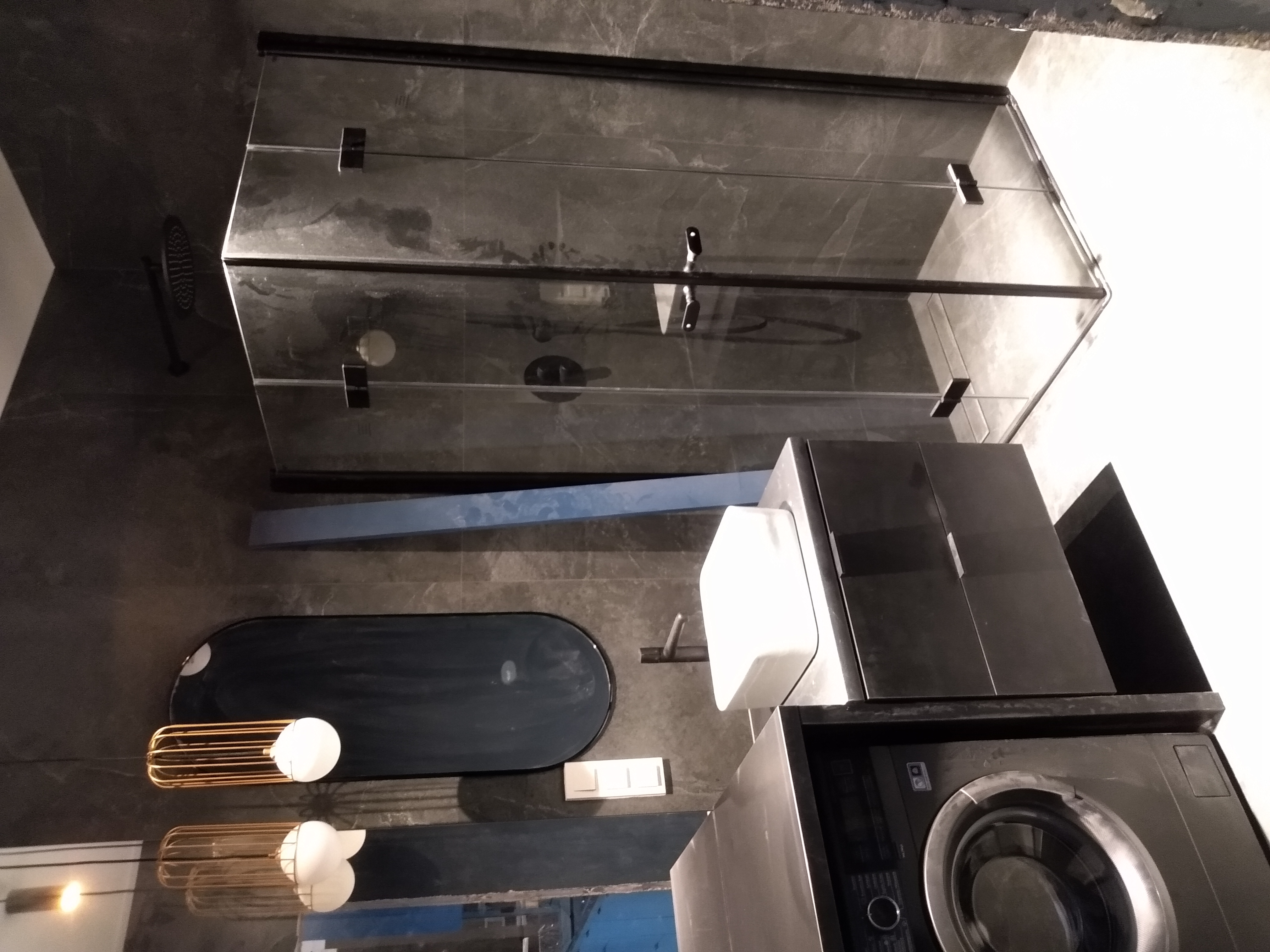 Nowoczesna łazienka w odcieniach szarości z kabiną prysznicową typu walk-in z czarnymi profilami, owalną umywalką, czarną szafką z blatem, pralką oraz dekoracyjnymi lampami wiszącymi z metalowymi...