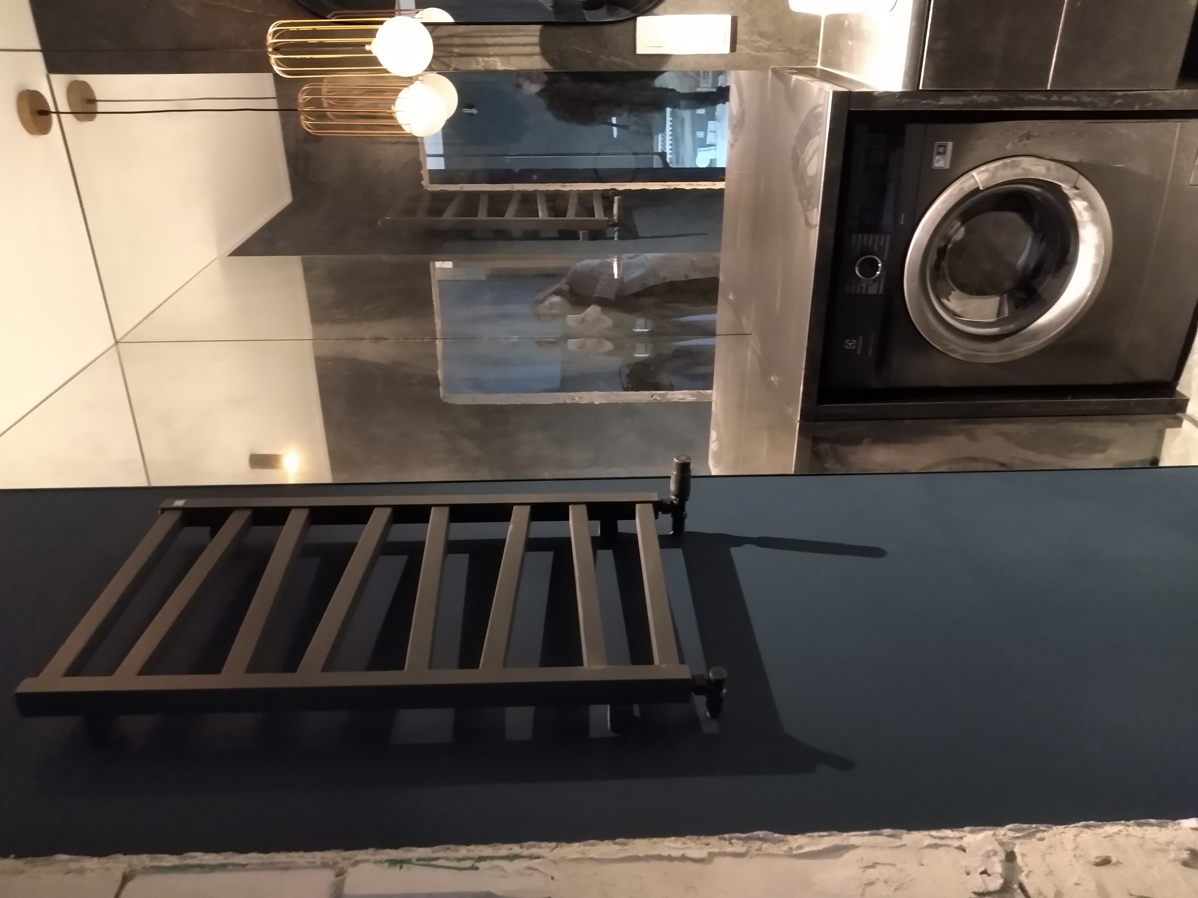 Szary grzejnik drabinkowy na ciemnoniebieskiej ścianie, z odbiciem łazienki w lustrzanej powierzchni, w tym pralka i oświetlenie.