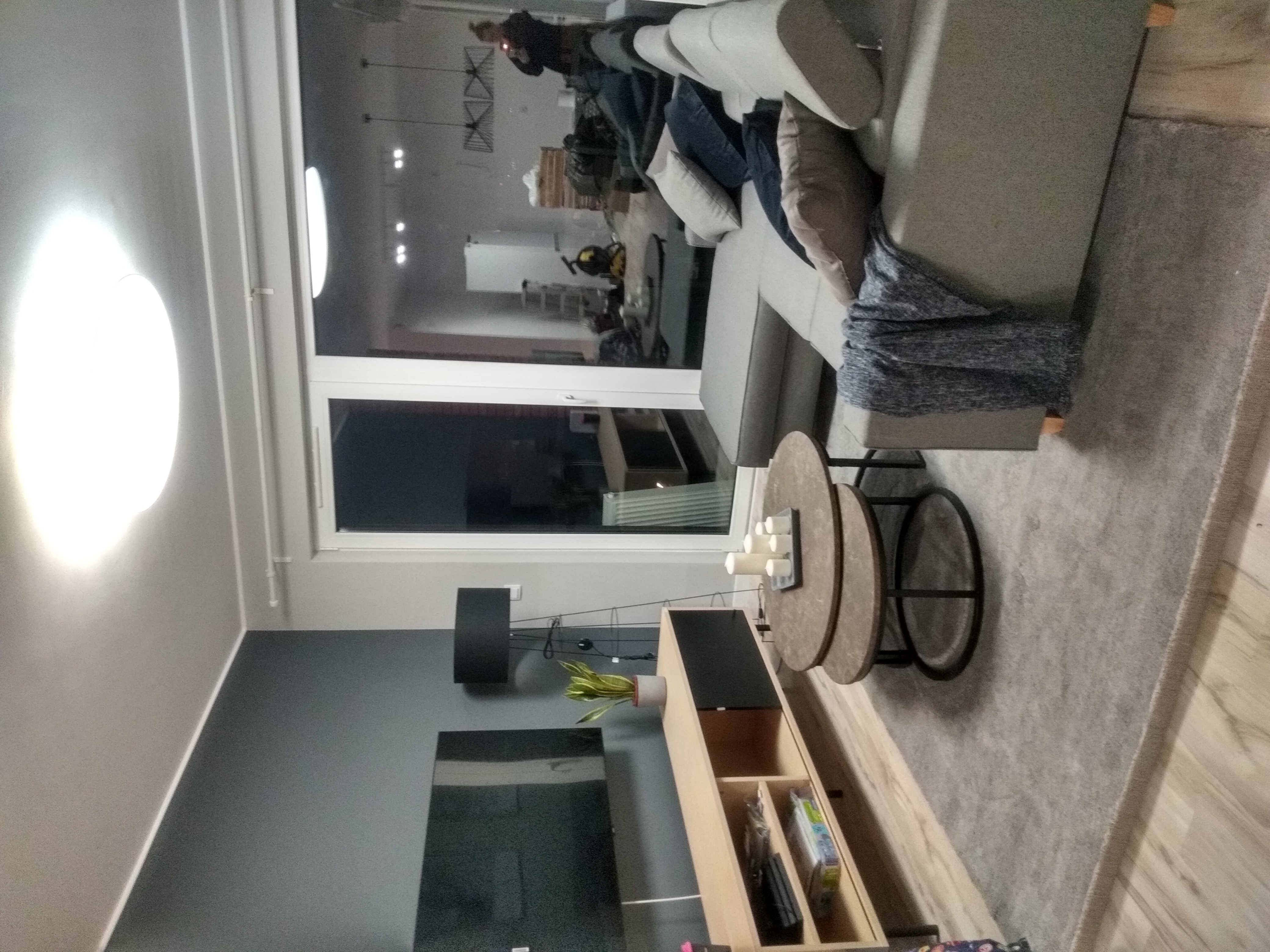 Nowoczesny salon z szarym narożnikiem, okrągłym stolikiem kawowym z dekoracyjnymi świecami, oraz szafką RTV z rośliną w doniczce, widok z boku, z odbiciem osoby w oknie.