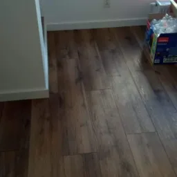 Ułożone panele podłogowe w odcieniach brązu w mieszkaniu we Wrocławiu, widoczna ściana z gniazdkiem elektrycznym oraz karton z akcesoriami do sprzątania.