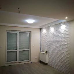 Pokój po remoncie z białą ceglaną ścianą, drewnianą podłogą, grzejnikiem i oknem balkonowym, widoczne ślady prac wykończeniowych.