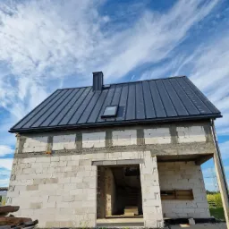 Surowy budynek w trakcie budowy z nowym, ciemnym dachem z blachy na tle błękitnego nieba z chmurami.