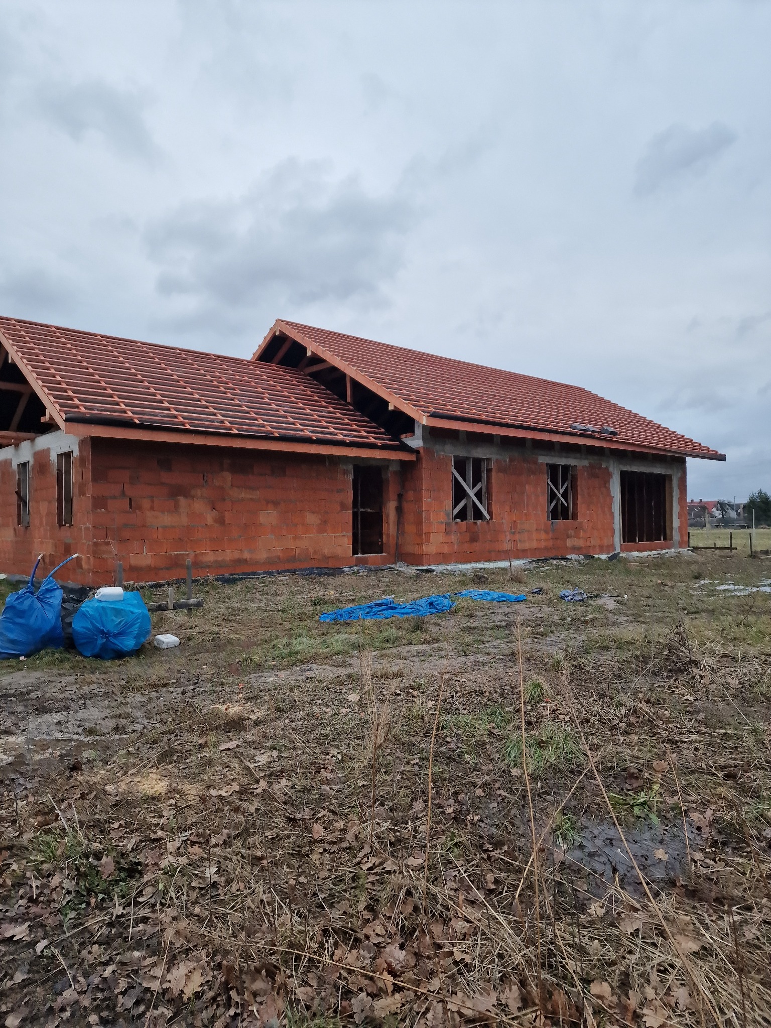 Dom w trakcie budowy z częściowo ukończonym dachem z czerwonej dachówki, widoczne krokwie i łaty, ściany z czerwonej cegły, okna zabite deskami, na pierwszym planie worki z materiałami budowlanymi...