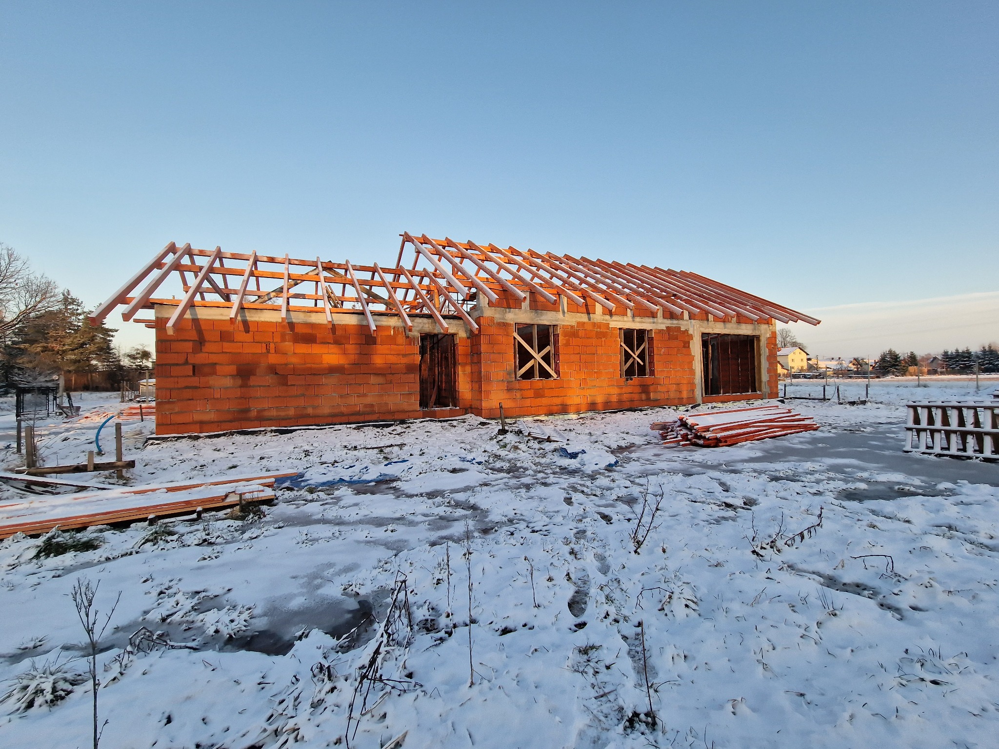 Surowy budynek z czerwonej cegły w zimowej scenerii, z częściowo zamontowaną drewnianą konstrukcją dachu, pokrytą lekkim śniegiem.