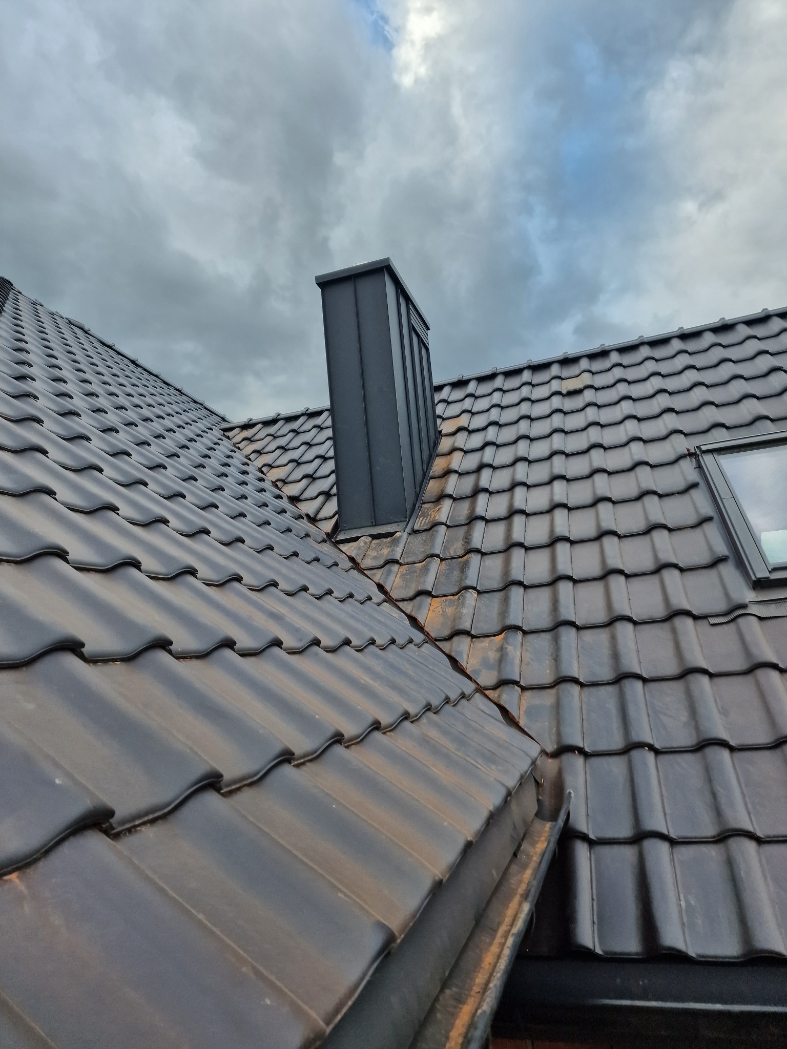 Nowoczesny dach z ciemnej dachówki ceramicznej z oknem dachowym i kominem, widok z góry na tle pochmurnego nieba.