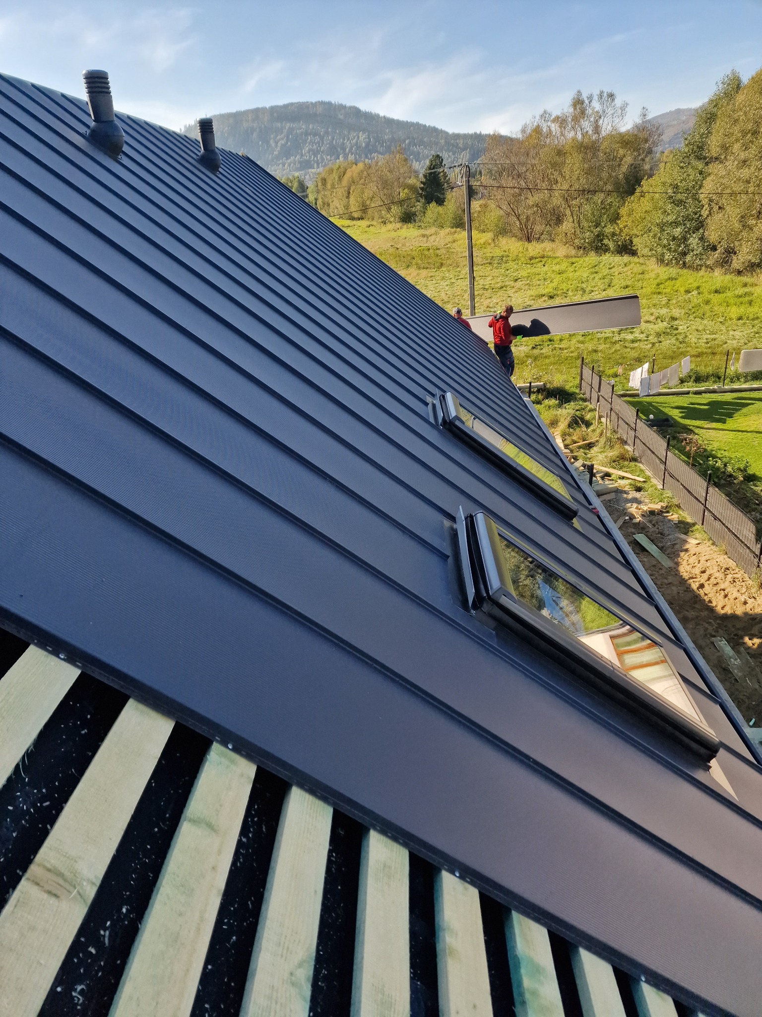 Montaż ciemnoszarej blachy na dachu z dwoma oknami dachowymi, widok na góry w tle, praca dekarzy w trakcie układania pokrycia.