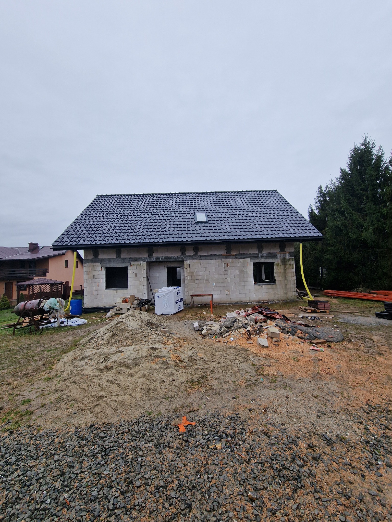 Surowy stan domu jednorodzinnego z czarną dachówką, widoczne bloczki betonowe, okna bez ram, rynna spustowa, materiały budowlane i gruz na posesji