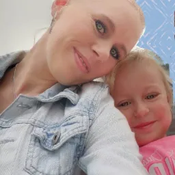 Portret uśmiechniętej kobiety w jeansowej kurtce obejmującej małą dziewczynkę w różowej koszulce.