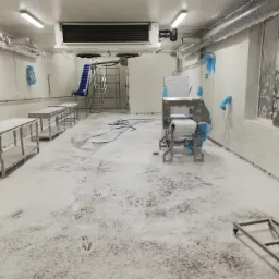 Wnętrze chłodni przemysłowej pokryte warstwą szronu i lodu, widoczne urządzenia chłodnicze, metalowe stoły, schody z taśmociągiem i ślady stóp na podłodze.