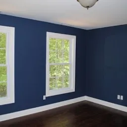 Pusty pokój z ciemnoniebieskimi ścianami, białymi listwami przypodłogowymi i ramami okiennymi oraz ciemną drewnianą podłogą. Widoczne dwa okna z widokiem na zieleń drzew na zewnątrz.