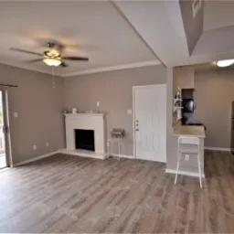 Jasne, przestronne wnętrze salonu z parkietem, kominkiem i otwartą kuchnią. Ściany w odcieniach beżu, białe drzwi i obudowa kominka. Widoczny fragment balkonu z drewnianą balustradą.