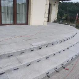 Wyłożony szarymi płytami kamiennymi taras z zaokrąglonymi schodami w trakcie budowy, widoczne elementy mocujące i fragment czerwonego kabla.