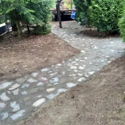 Ścieżka z nieregularnych kamieni prowadząca przez ogród, otoczona zielenią i drzewami iglastymi, z widocznym samochodem w oddali.