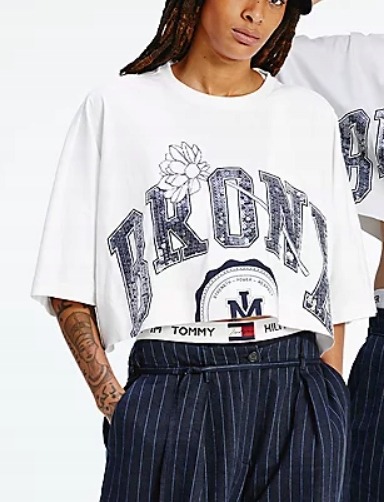 Krótki, biały T-shirt z nadrukiem 'BRONX' i motywem kwiatowym, spod którego wystaje pasek z logo Tommy Hilfiger, zestawiony z granatowymi spodniami w prążki. Modelka z tatuażem na przedramieniu.