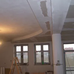 Niedokończony podwieszany sufit z elementami gipsowymi, widoczne łączenia i szpachlowanie, w tle okna i drabina malarska.