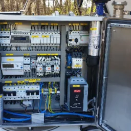 Otwarta, nierdzewna szafa sterownicza IP68 z regulatorem mocy biernej, softstarterem Danfoss, bezpiecznikami EATON i kondensatorami ETI, zainstalowana na zewnątrz, prawdopodobnie dla pompy głębinowej.