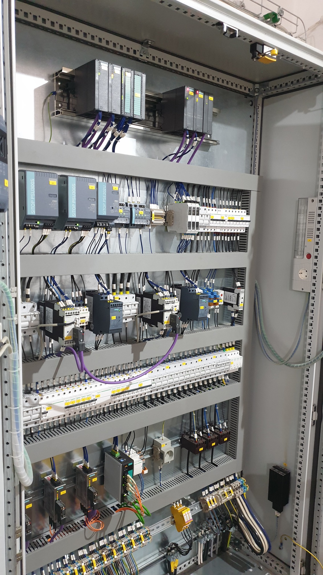 Szara rozdzielnica elektryczna z zamontowanymi sterownikami Siemens S7 i licznymi przewodami w różnych kolorach, zainstalowana wewnątrz metalowej szafy sterowniczej. Widoczne moduły zasilające...