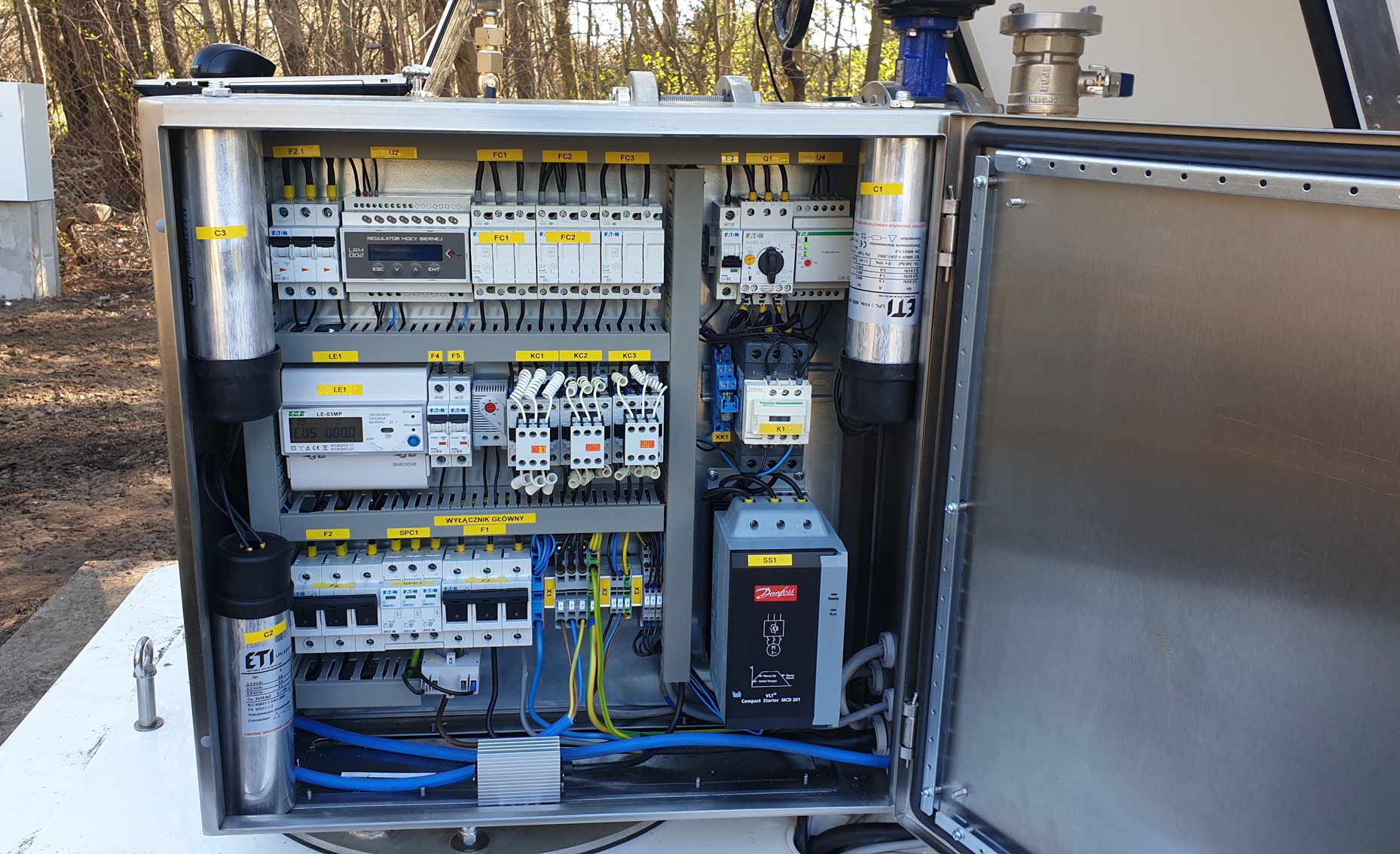 Otwarta, nierdzewna szafa sterownicza IP68 z regulatorem mocy biernej, softstarterem Danfoss, bezpiecznikami EATON i kondensatorami ETI, zainstalowana na zewnątrz, prawdopodobnie dla pompy głębinowej.