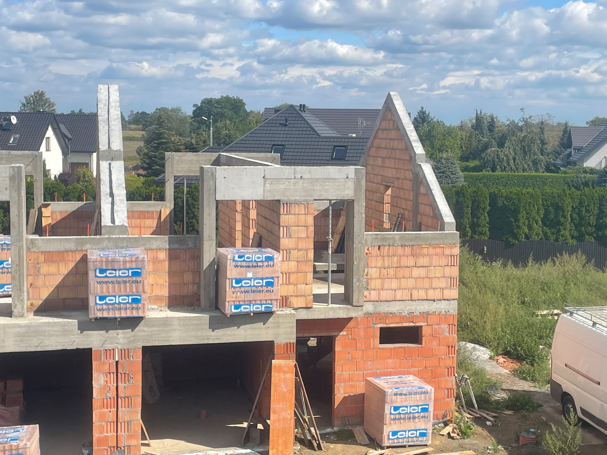 Wznoszenie ścian domu z czerwonej cegły, widoczne betonowe elementy konstrukcyjne, palety cegieł na budynku, w tle zabudowa jednorodzinna i zieleń.