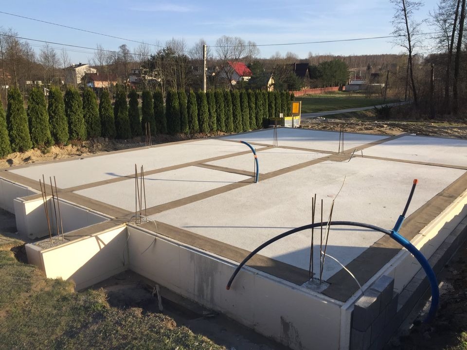 Zalana płyta fundamentowa z wystającymi prętami zbrojeniowymi i częściowo wymurowanymi ścianami fundamentowymi z bloczków betonowych, widoczne niebieskie rury instalacyjne, w tle rząd tuj...