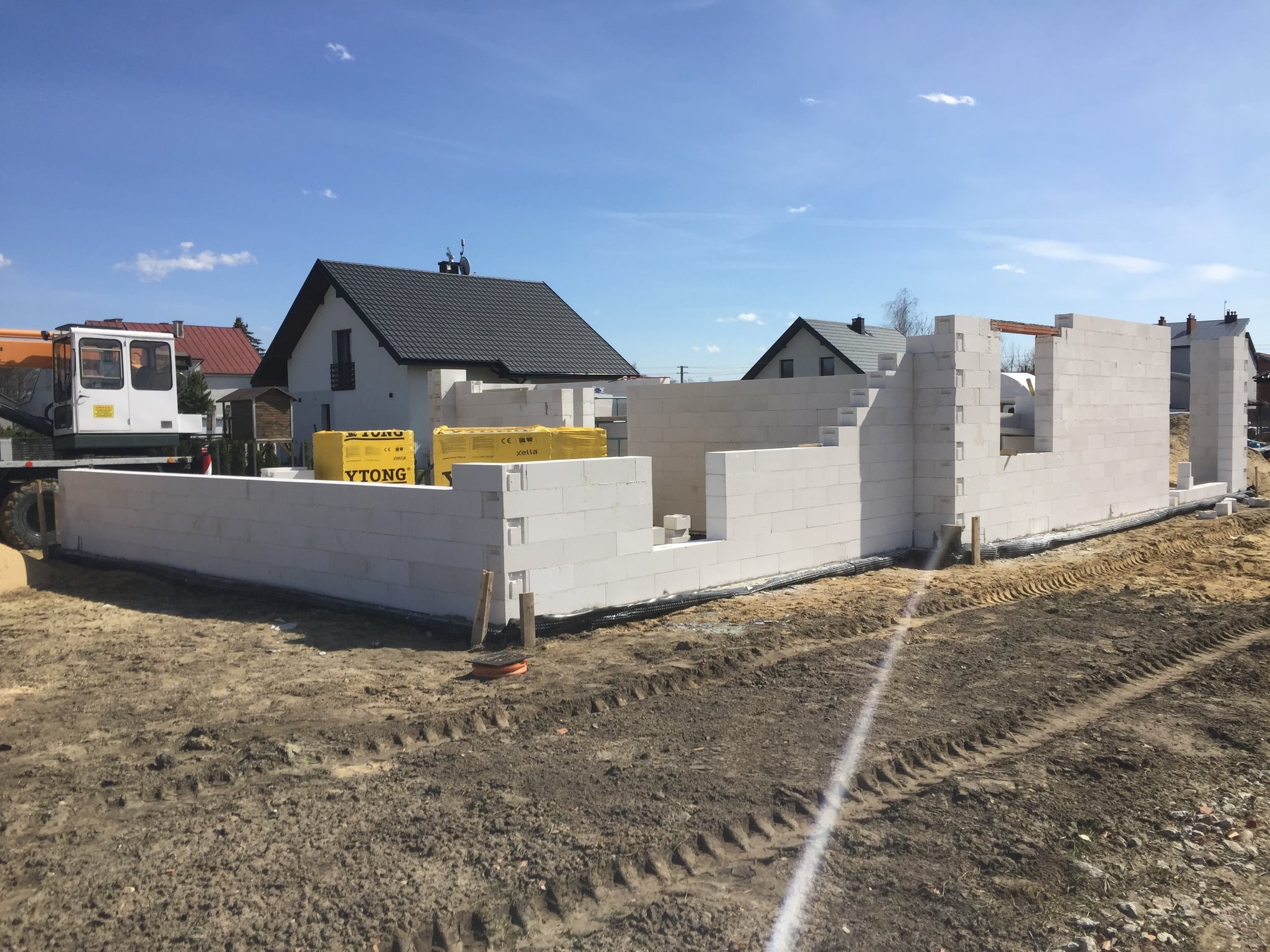 Budowa domu z bloczków Ytong: widoczne fundamenty, pierwsze ściany, palety z bloczkami, koparka w tle, domy sąsiadów w oddali.