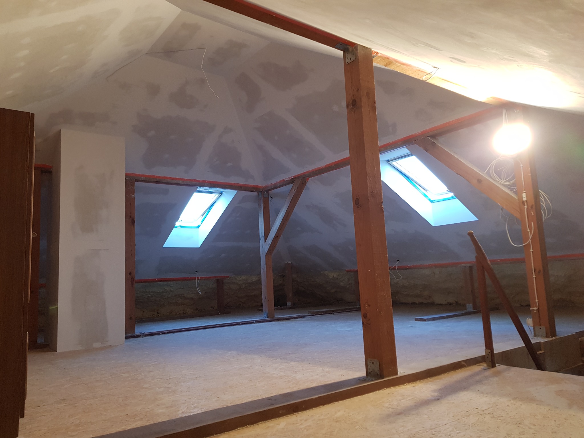 Poddasze w trakcie adaptacji: surowe drewniane belki konstrukcyjne, dwa okna dachowe wpuszczające jasne światło, ściany częściowo pokryte gładzią szpachlową, widoczna izolacja na ścianach...