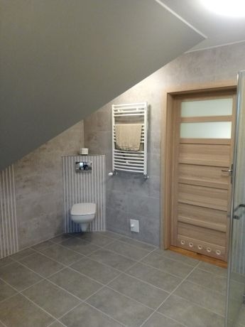 Ukośna łazienka na poddaszu z szarymi płytkami, wiszącą toaletą, grzejnikiem drabinkowym z ręcznikiem i drewnianymi drzwiami z przeszkleniami.