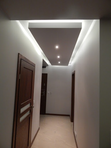 Korytarz z podwieszanym sufitem z oświetleniem LED i punktowymi lampami, widoczne drzwi z drewnianą okleiną i przeszklonym panelem.