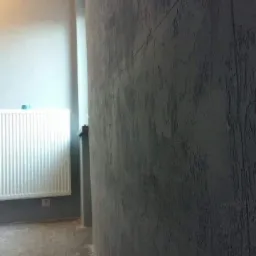 Szara ściana z wyraźną fakturą, widoczny fragment grzejnika i podłoga, surowe wykończenie wnętrza.