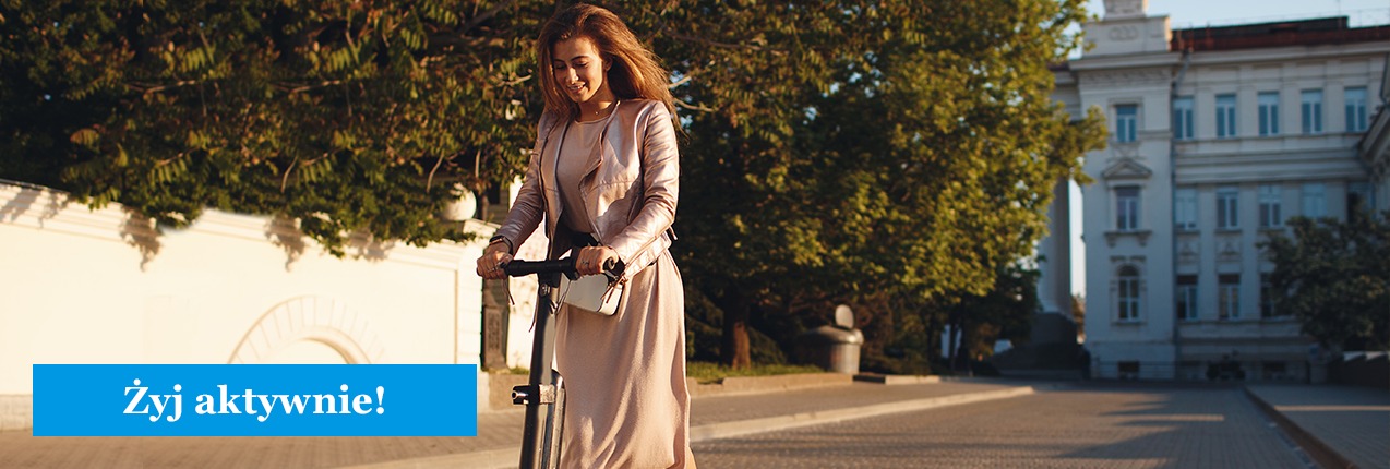 Uśmiechnięta kobieta w eleganckim stroju jedzie na hulajnodze elektrycznej po brukowanej ulicy w słoneczny dzień, w tle budynek z klasyczną architekturą i drzewa.