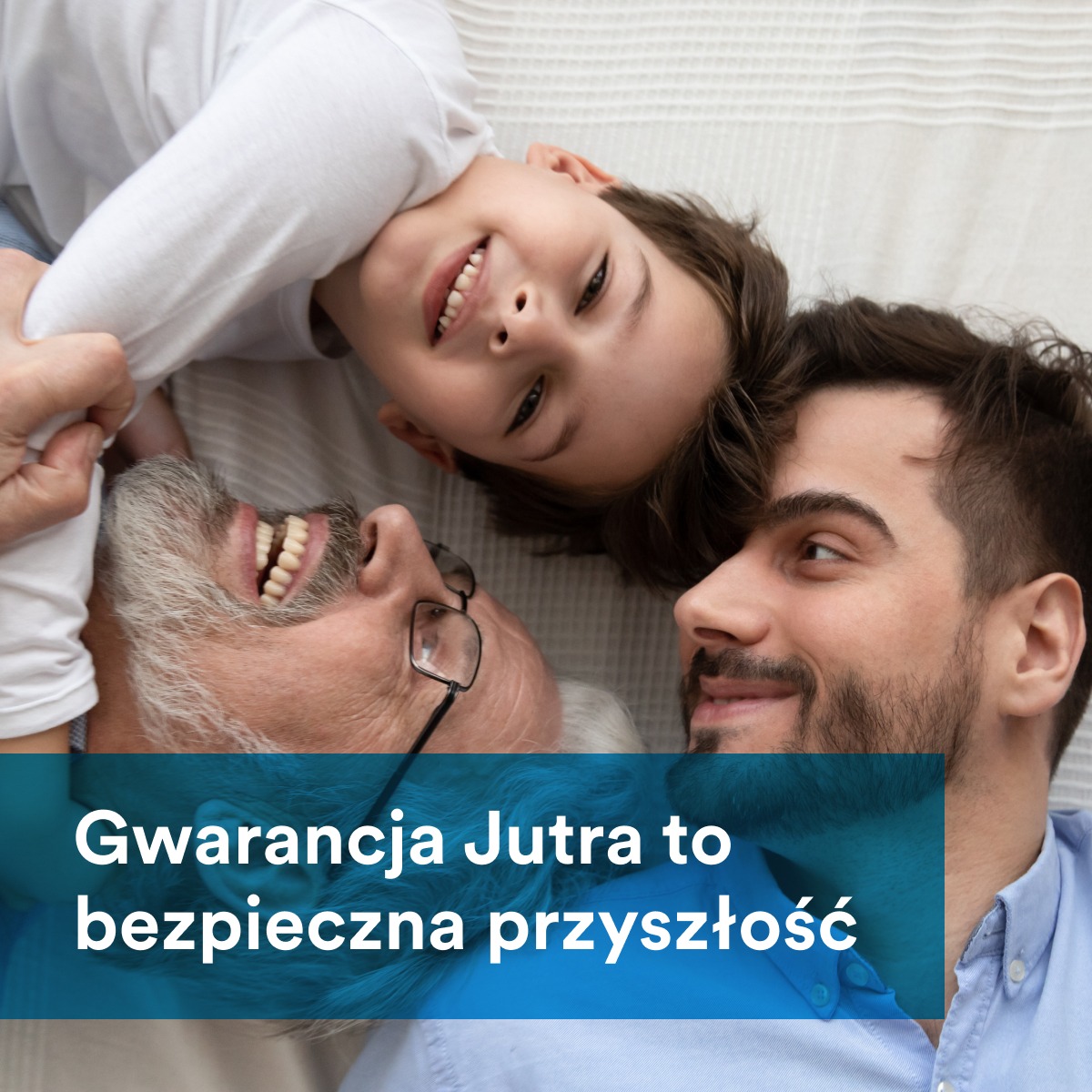 Trzy pokolenia mężczyzn: dziadek w okularach, ojciec z brodą i uśmiechnięty syn leżący na jasnym tle, patrzący w górę.
