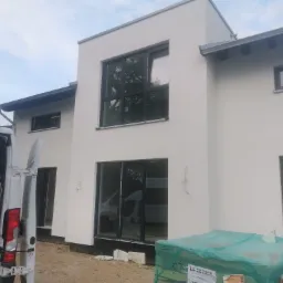 Nowoczesny dom z jasną, gładką elewacją, widoczne okna z czarnymi ramami, częściowo otwarty samochód dostawczy z narzędziami i materiałami w tle.