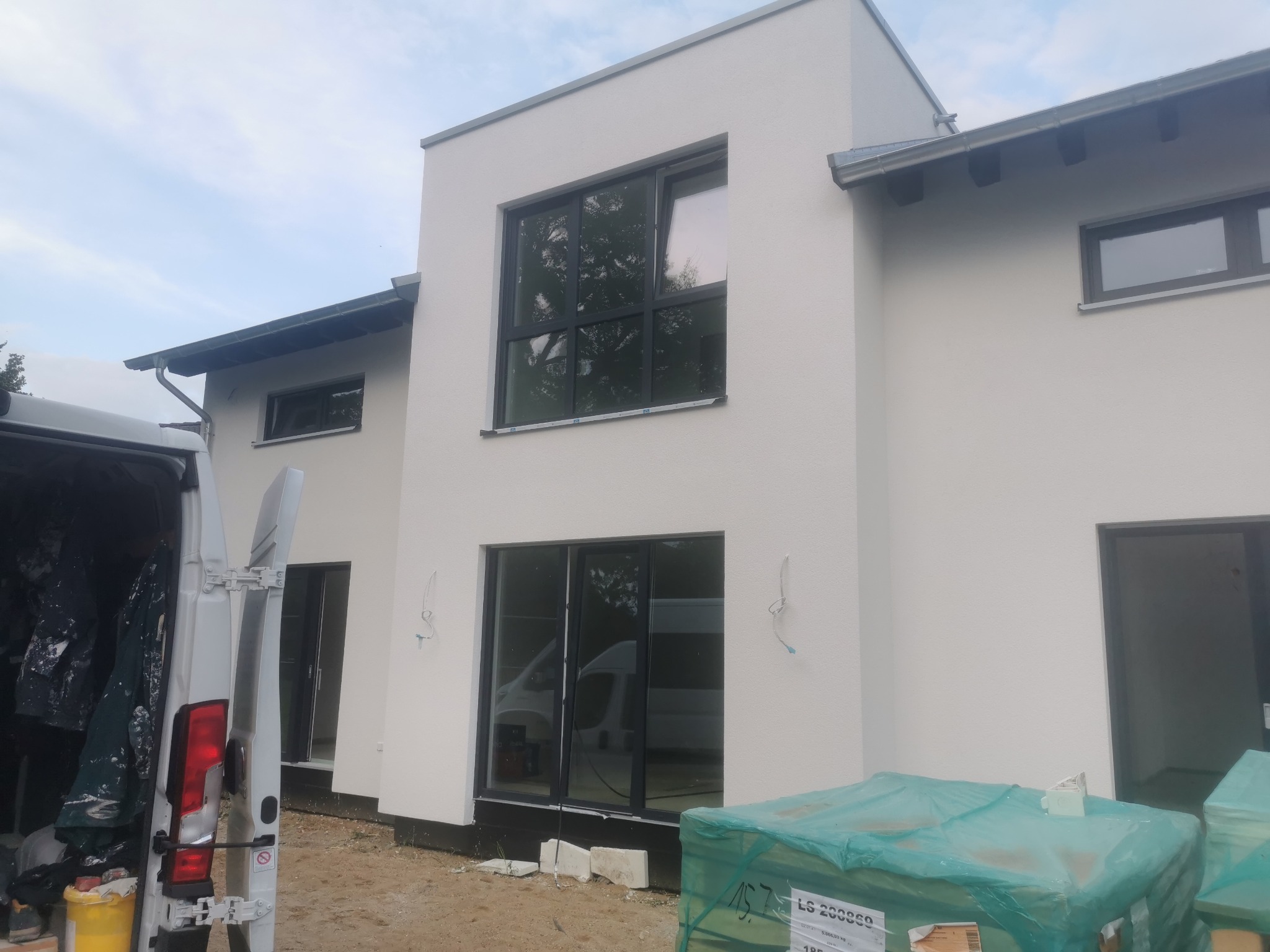 Nowoczesny dom z jasną, gładką elewacją, widoczne okna z czarnymi ramami, częściowo otwarty samochód dostawczy z narzędziami i materiałami w tle.