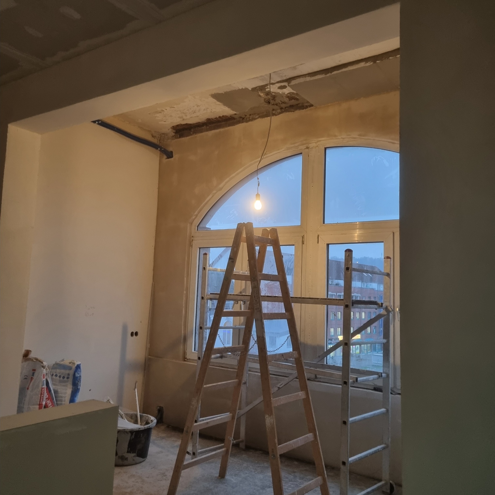 Wnętrze w trakcie remontu z drewnianą drabiną na tle okna z łukiem, widoczne worki z zaprawą i surowy sufit z widocznymi elementami konstrukcyjnymi.