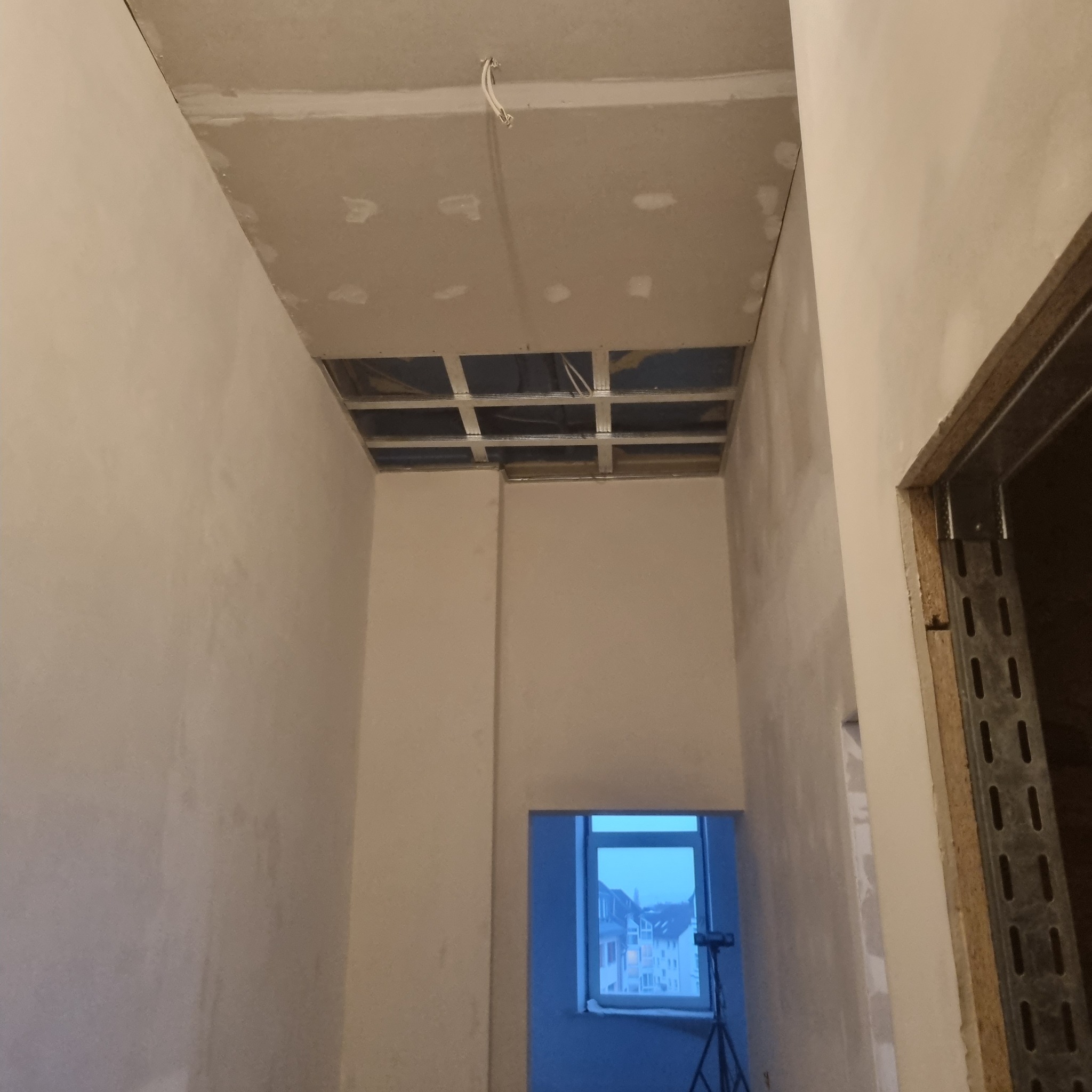 Wnętrze w trakcie prac wykończeniowych; widoczny sufit podwieszany z odsłoniętą metalową konstrukcją, ściany po gruntowaniu z widocznymi punktowymi poprawkami gipsowymi, surowe ościeżnice metalowe,...